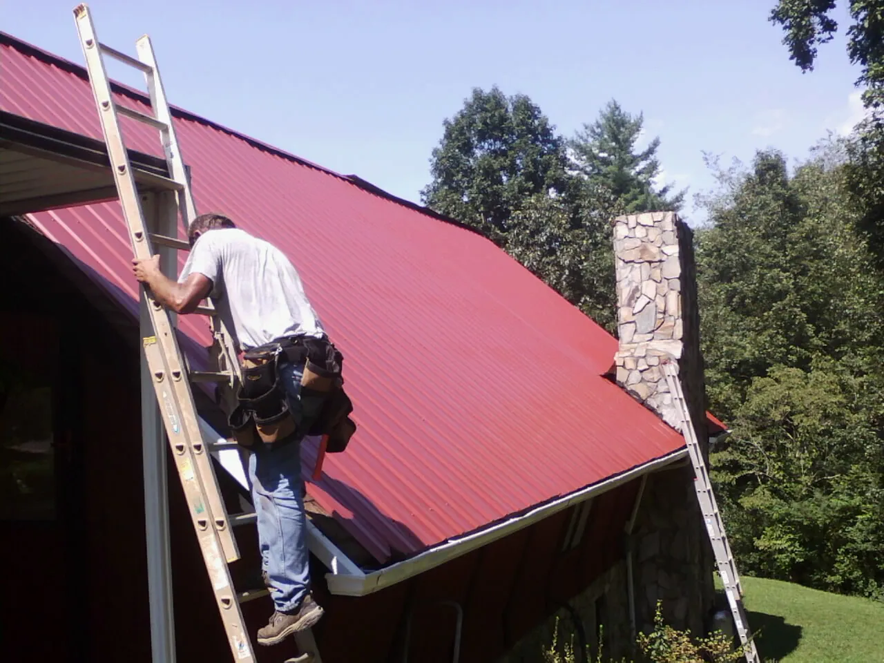 Roofer on a ladder beside a red metal roof and stone chimney — Soffit & Fascia Repair by Roofing Brothers in Keno